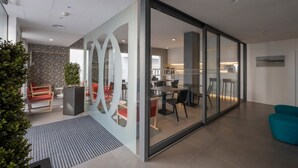 Espaço de escritório moderno com divisória de vidro, área de refeições com cadeiras vermelhas e uma cozinha ao fundo.