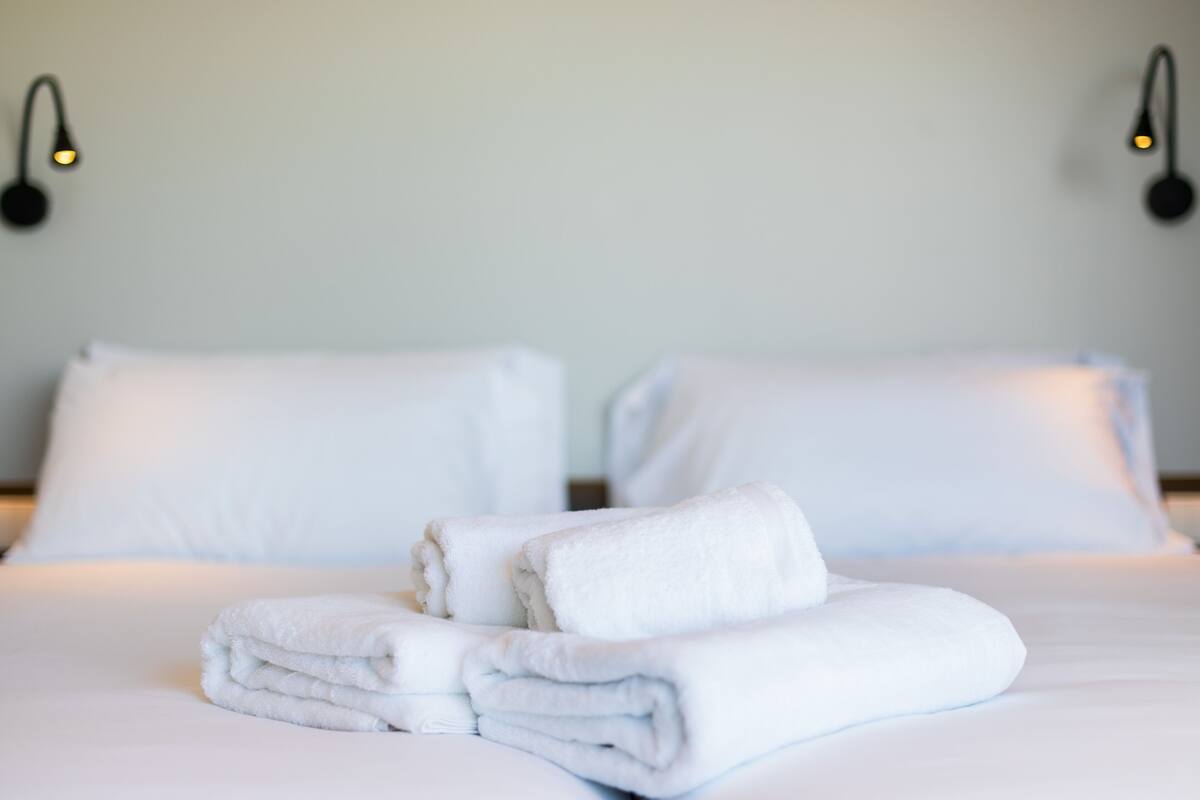 A neatly made bed with white linens and folded towels, two wall-mounted light fixtures, and a plain wall.