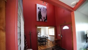 Una habitación con una pared roja, una puerta de madera, un perchero, una mesa de comedor con sillas y una obra de arte de vaca colgada del techo.