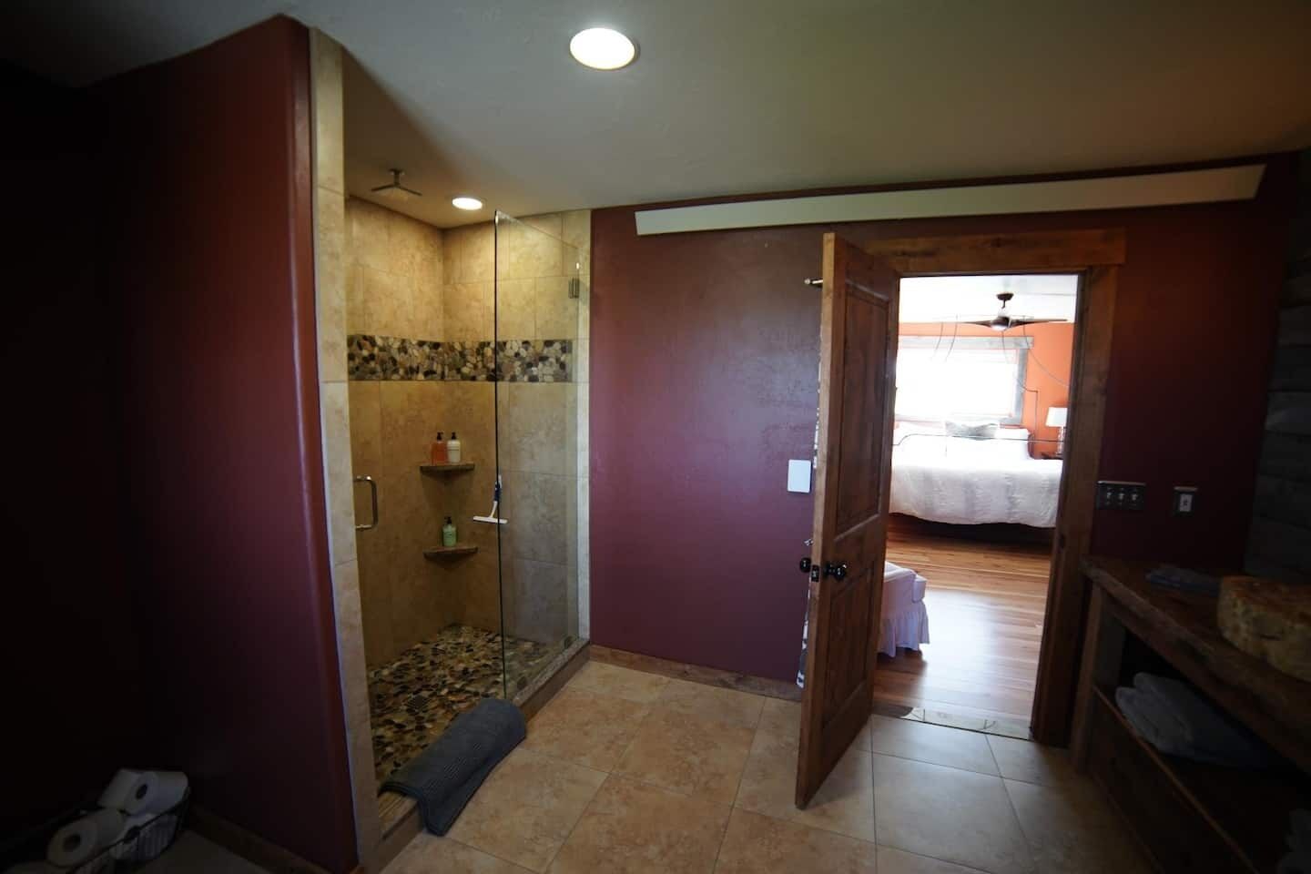 A bathroom with a glass-enclosed shower, a wooden door leading to a bedroom, and a tiled floor.