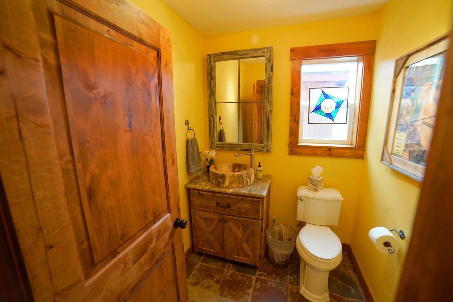 Ein Badezimmer mit Holztür, Waschbecken mit Steinbecken, Toilette und Fenster mit Buntglasbild.