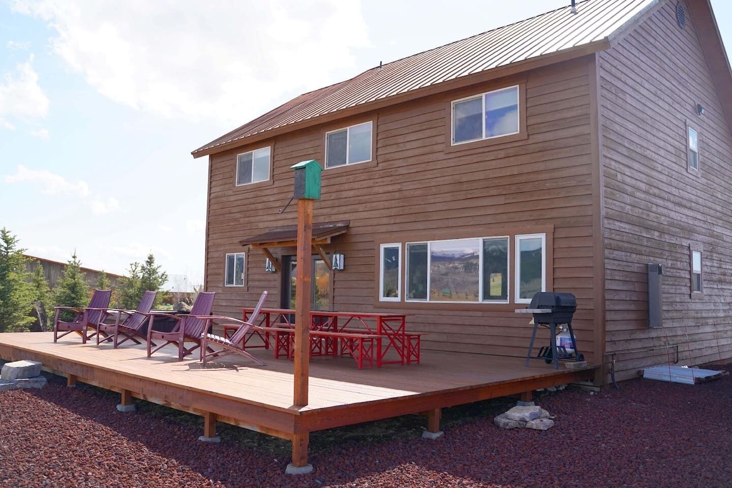 A two-story wooden house with a deck, outdoor seating, and a grill.