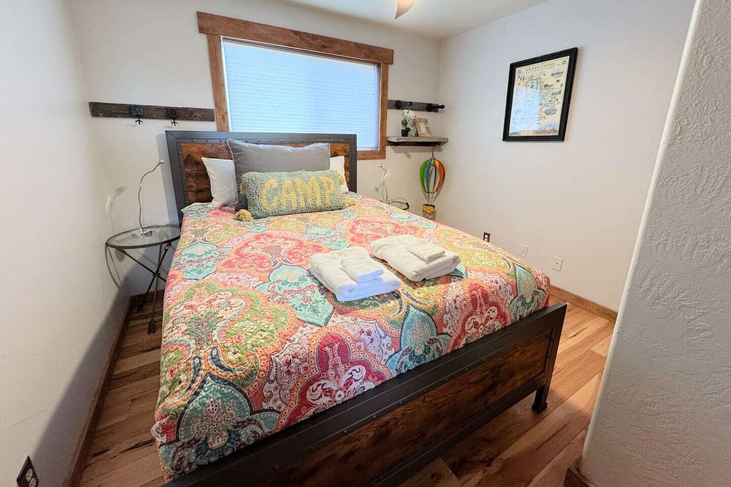 Une chambre à coucher avec un cadre de lit en bois, une couvre-lit aux motifs colorés, une fenêtre avec des stores, une petite table de chevet et une étagère murale sur laquelle est posée une photo encadrée.