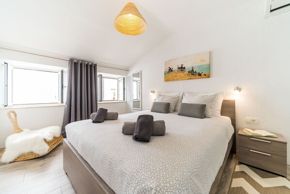 A bedroom with a large bed, a bedside table with a lamp, a wall-mounted painting, and a window with curtains.