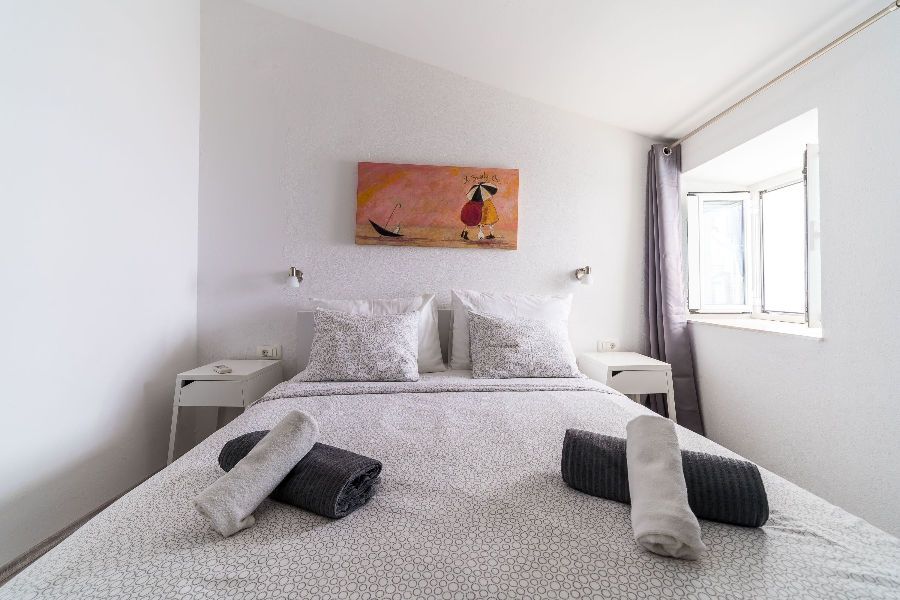 A neatly made bed with two pillows, a bedside table, a wall painting, and a window with curtains.