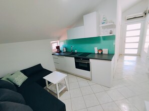 A compact kitchen with white cabinets, a black countertop, and a green backsplash. There is a small white table and a black sofa with a green pillow.