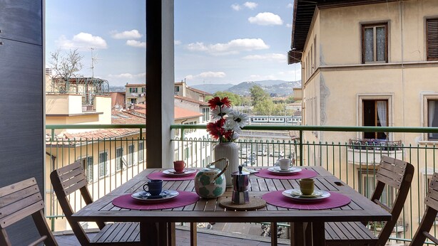 A balcony with a table set for two, a vase with flowers, and a view of buildings and mountains. - Mamo Florence - Binario 9 Apartment (Florence)