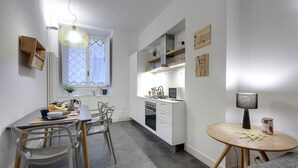A modern kitchen with white cabinetry, a dining area with a table and chairs, and a small round table with a lamp and jar. - Mamo Florence - Costa San Giorgio Apartment (Florence)
