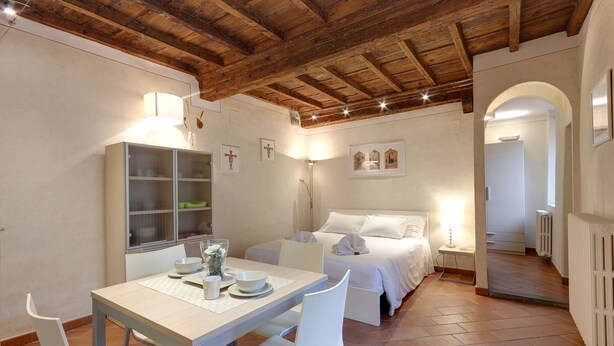 A room with a wooden ceiling, a bed with white bedding, a dining table with chairs, and a cabinet with glass doors. - Mamo Florence - Cupolone Apartment (Florence)