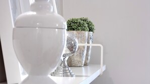 Una lámpara de pedestal blanca, dos jarrones decorativos con una planta y un estante blanco contra una pared lisa.