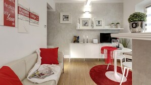Una sala de estar moderna con un sofá blanco, cojines rojos, un tapete rojo, una zona de comedor con una mesa y sillas blancas, y una cocina con un armario blanco y una planta en maceta.