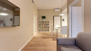 Een moderne woonkamer met een grijze bank, houten vloeren, een wit eetgedeelte met een tafel en stoelen, en een boekenkast met decoratieve objecten.