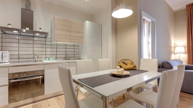 Een moderne keuken met een inbouwoven, een eethoek met tafel en stoelen, en een zithoek met een bank en een lamp.