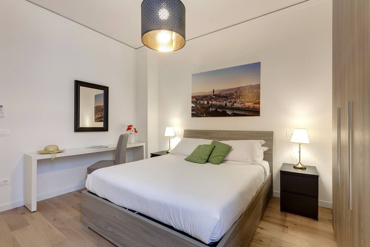 A neatly arranged bedroom with a bed, bedside tables, a nightstand, a wall-mounted lamp, and a cityscape picture.