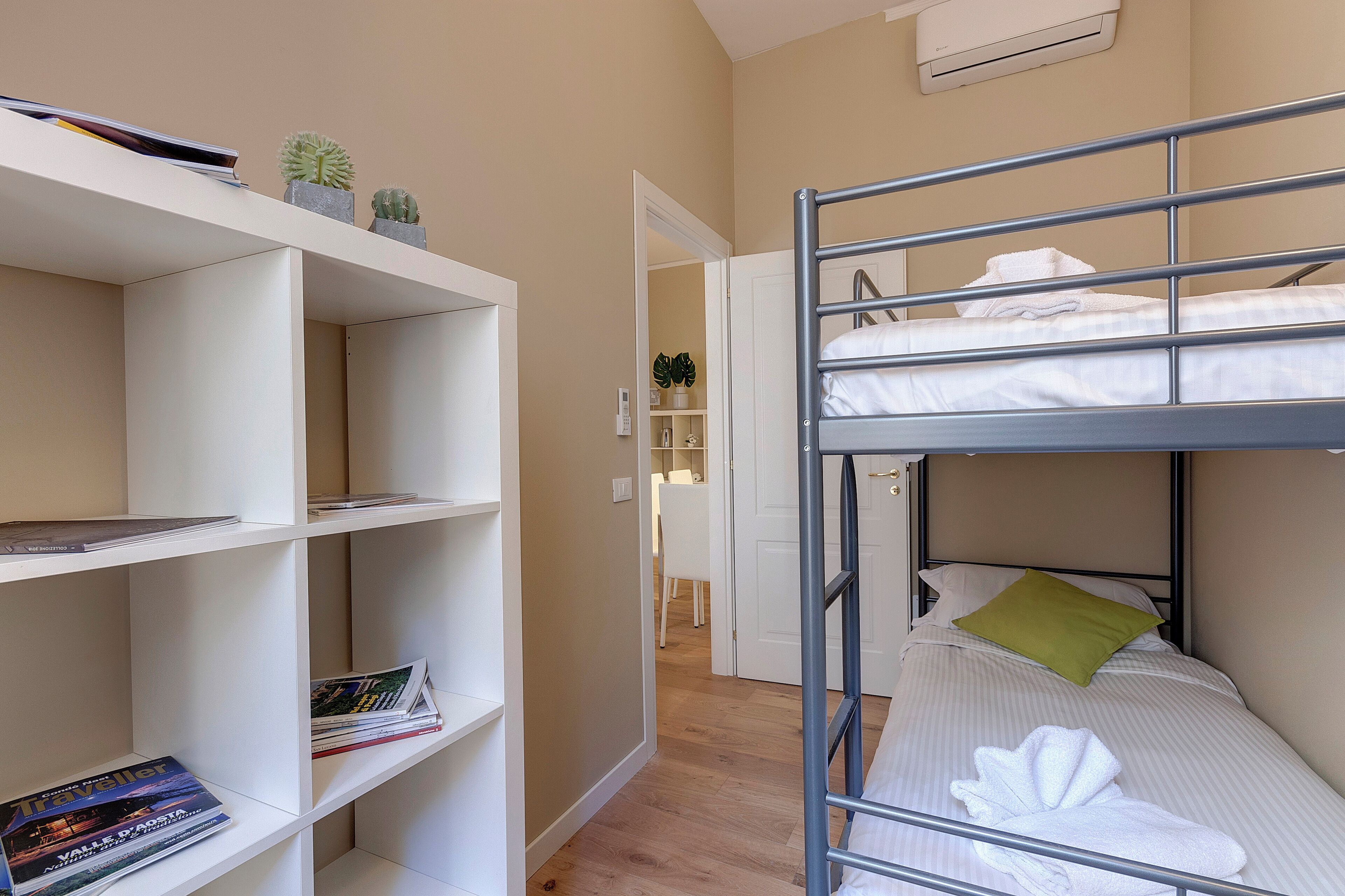 Un lit superposé avec un oreiller blanc et vert, une étagère blanche avec des livres et une petite plante, et une porte blanche donnant sur une autre pièce.