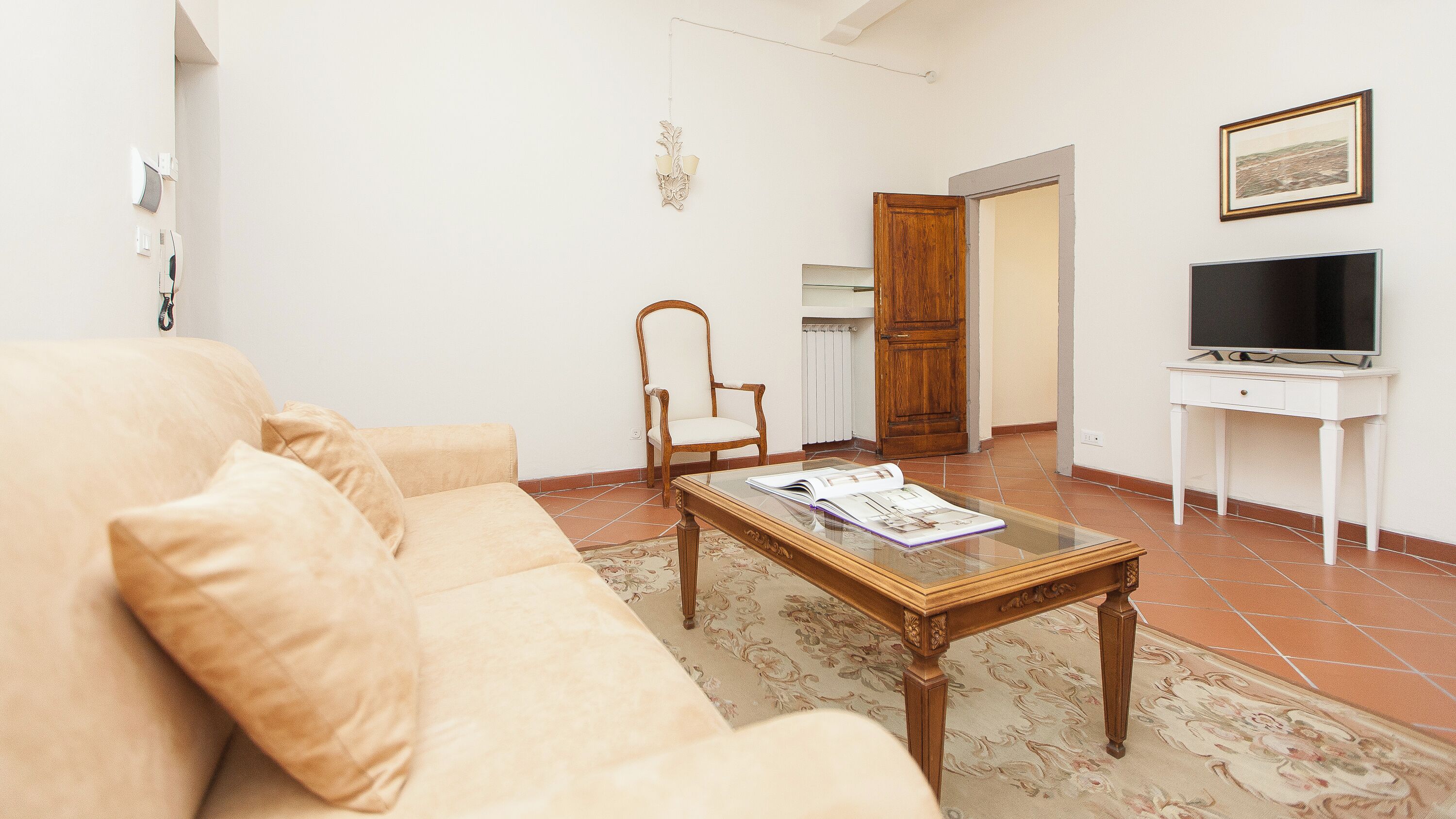 A living room with a beige sofa, a wooden chair, a glass coffee table with magazines, a white desk with a TV, and a wooden wardrobe.