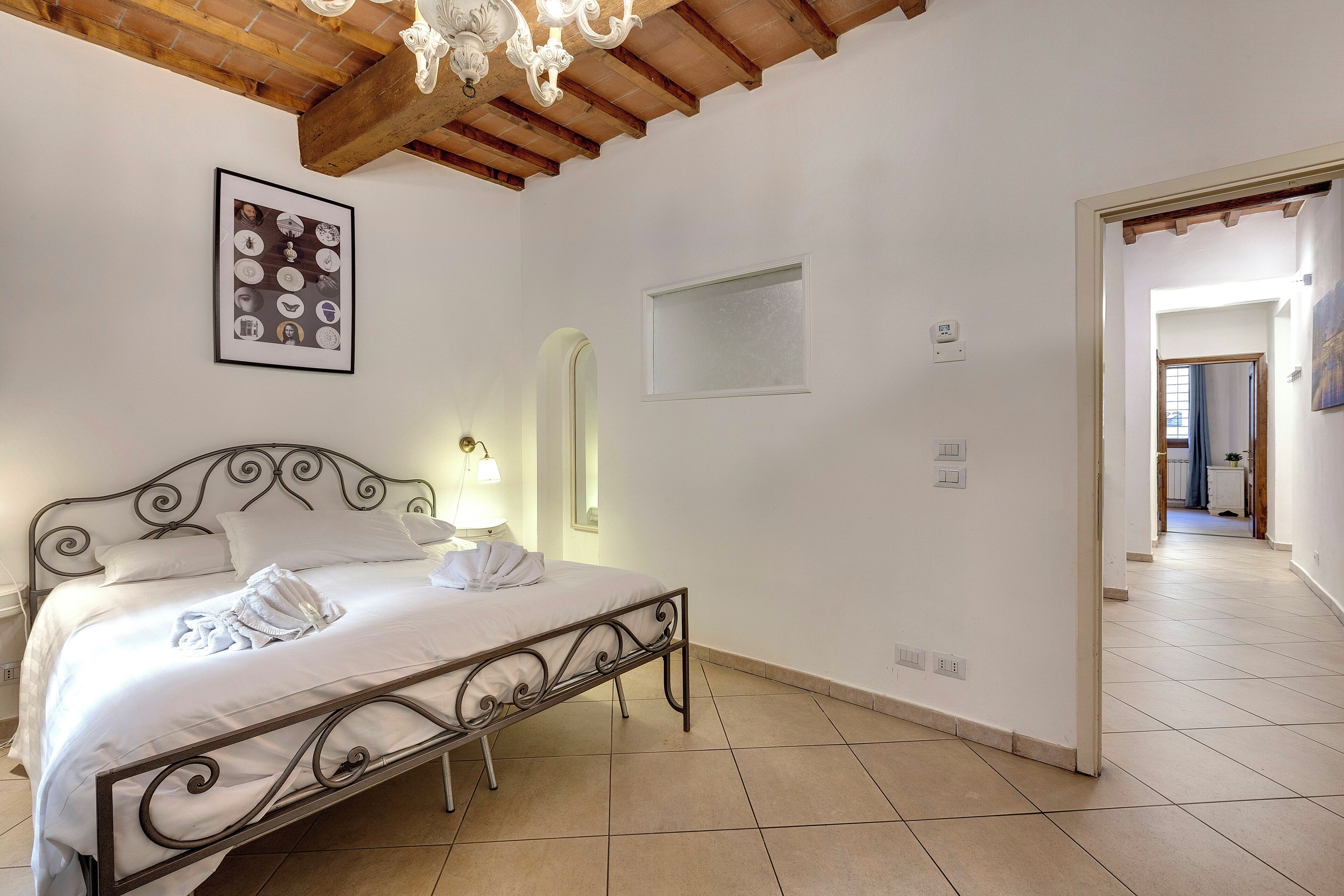 A bedroom with a bed, a chandelier, a framed picture, and a tiled floor.