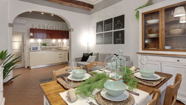 A dining area with a wooden table set for four, surrounded by chairs. There is a kitchen visible in the background with a 'KITCHEN' sign. - Mamo Florence - Ghibellina Apartment (Florence)