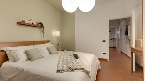 A bedroom with a large bed, a wooden headboard, a bedside table with a lamp, and a wall-mounted shelf with books and a plant. - Mamo Florence - Ghibellina Apartment (Florence)