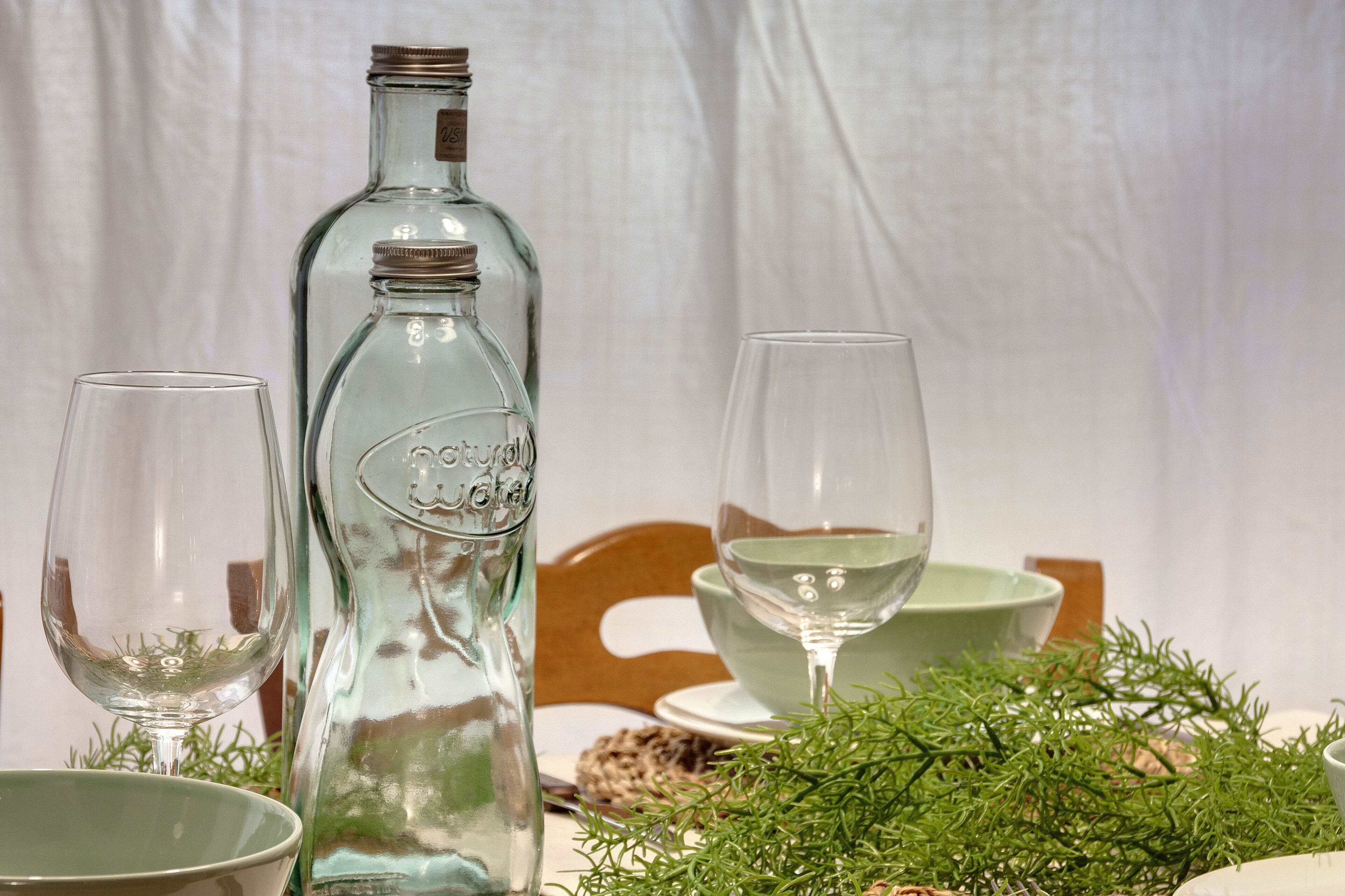 Une table mise avec une bouteille en verre transparent, deux verres à vin et un bol vert.