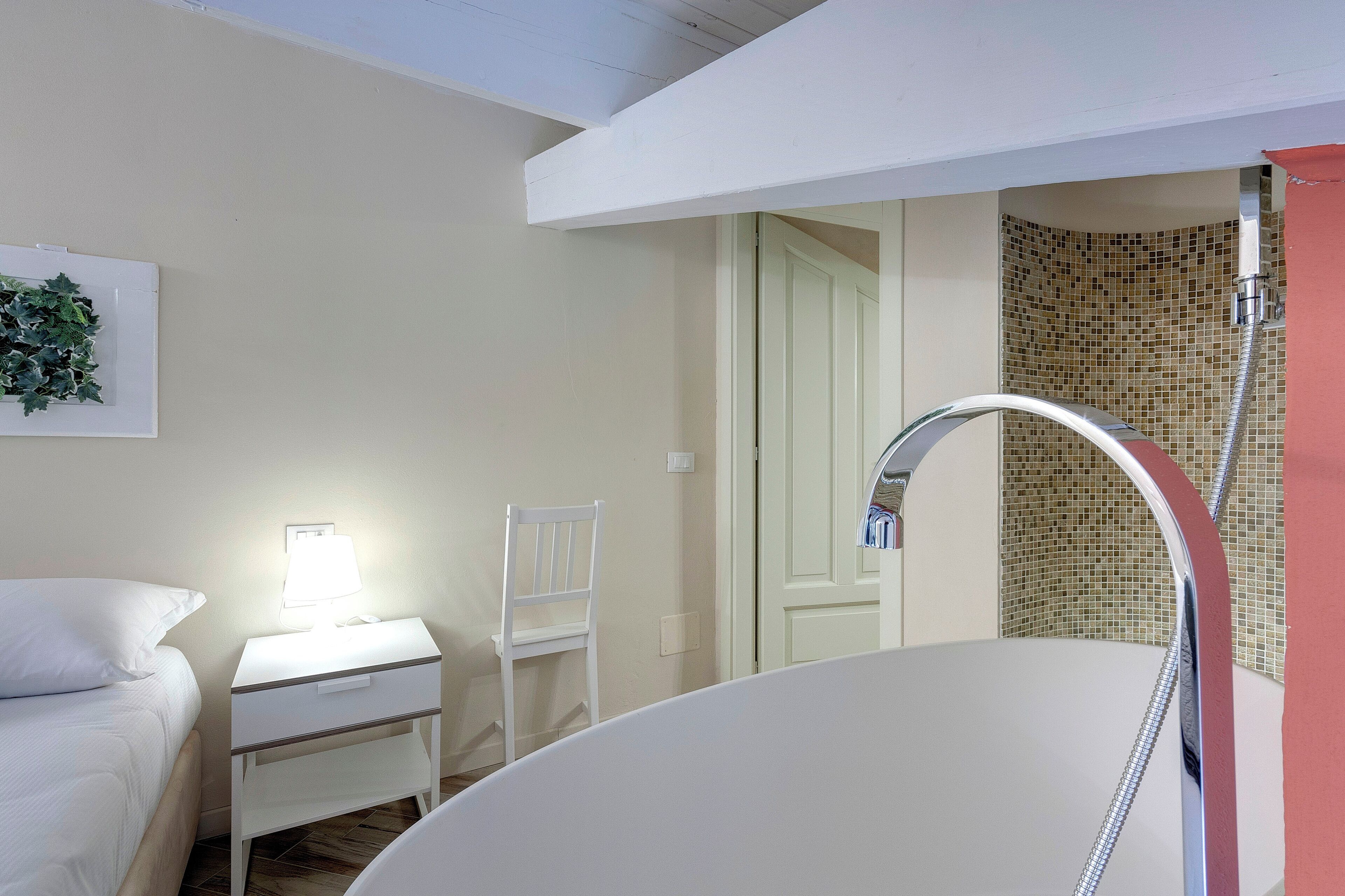 Une salle de bain avec une baignoire, une table de chevet blanche avec une lampe, une chaise blanche et un espace douche avec un mur carrelé.
