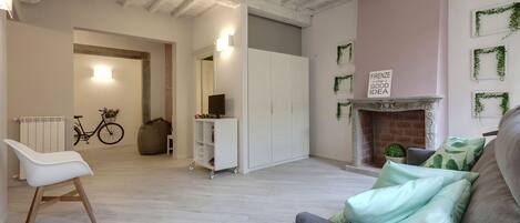 A spacious living room with a grey sofa, a white chair, and a white cabinet. A bicycle is parked in the background, and a sign on the wall reads "FIRENZE is always a good idea."