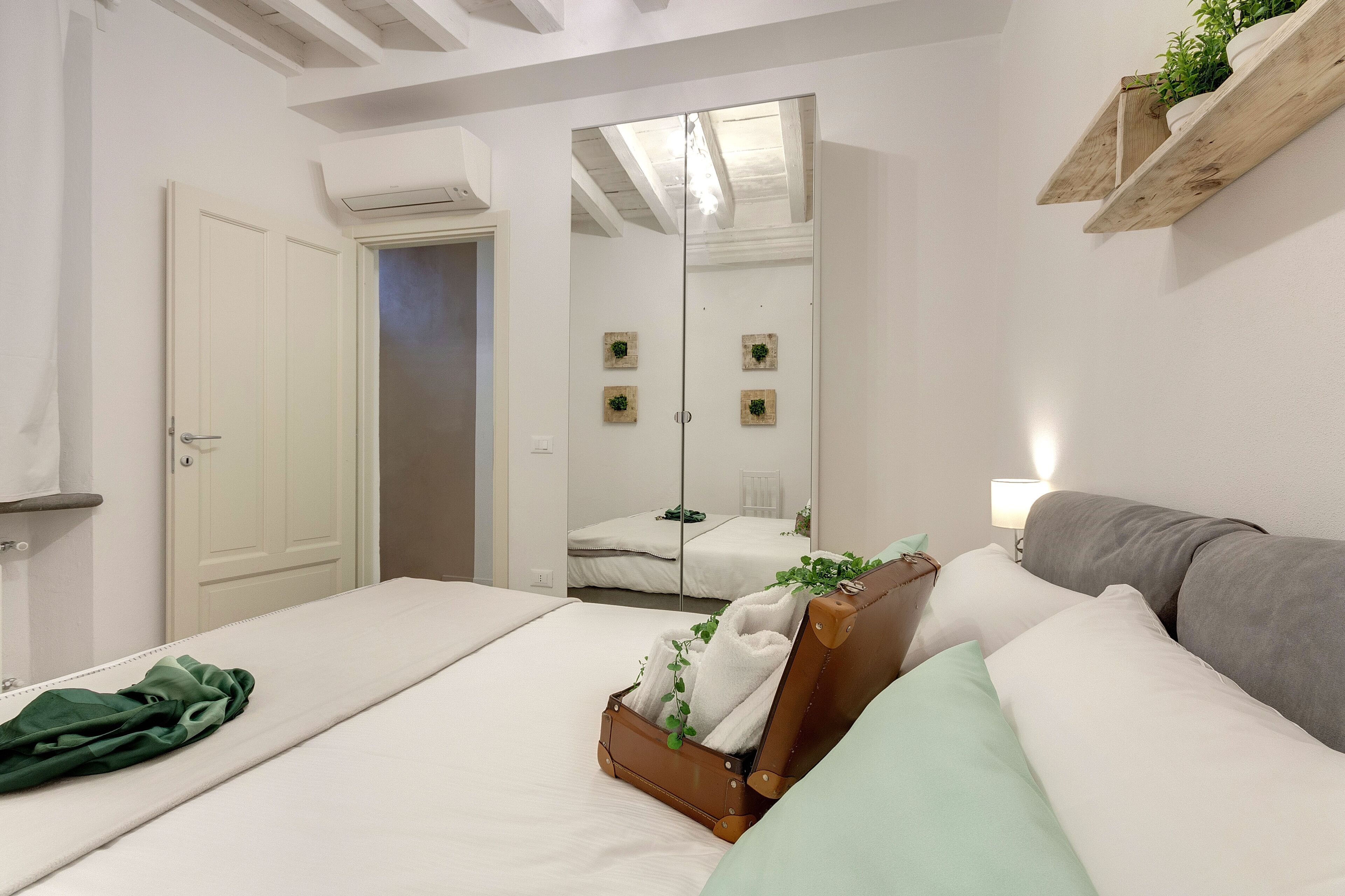 A bedroom with a bed, a suitcase, a green blanket, a grey sofa, and a mirror.