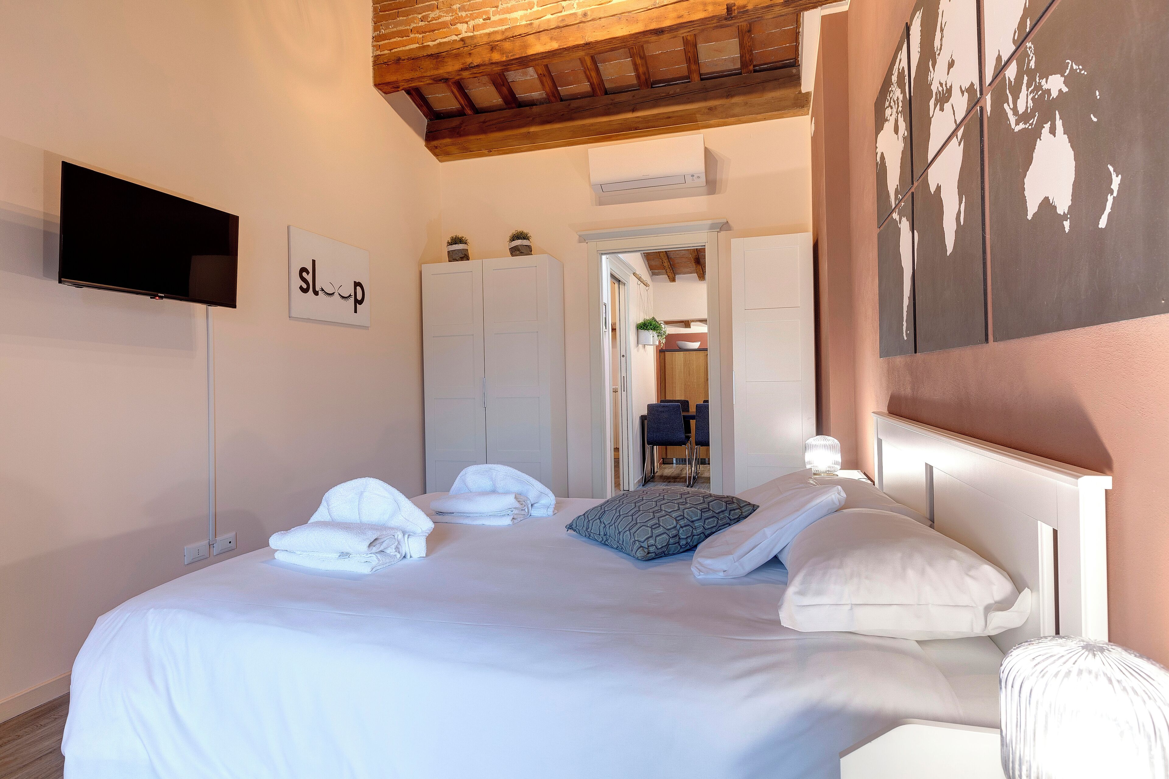 A neatly made bed with white linens and pillows, a flat-screen TV mounted on the wall, and a world map artwork on the wall.