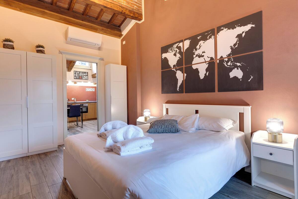 A neatly made bed with towels, a world map on the wall, and a kitchen area visible through an open door.