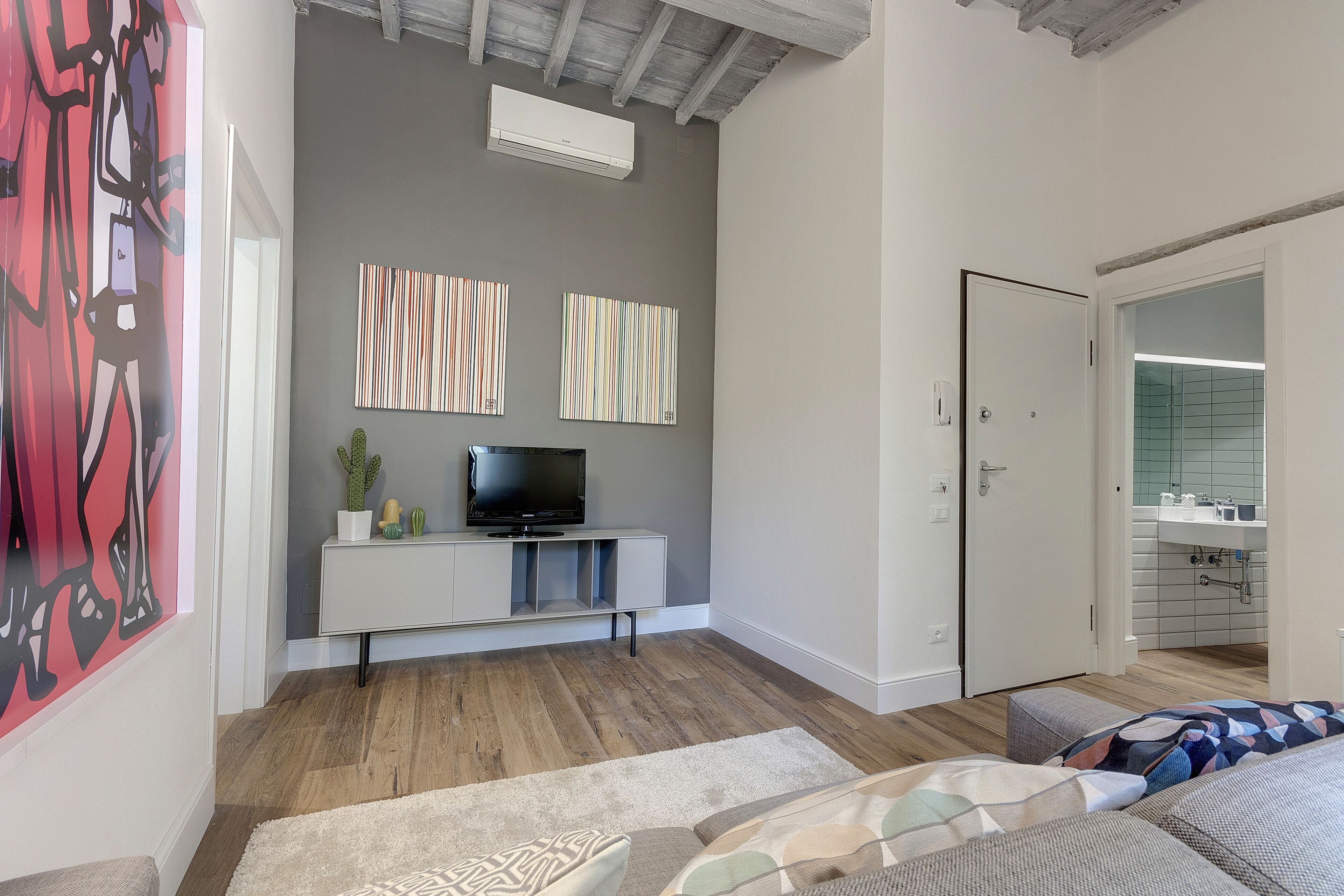 A modern living room with a grey wall, wooden flooring, a white entertainment unit with a flat-screen TV, and a door leading to a bathroom.