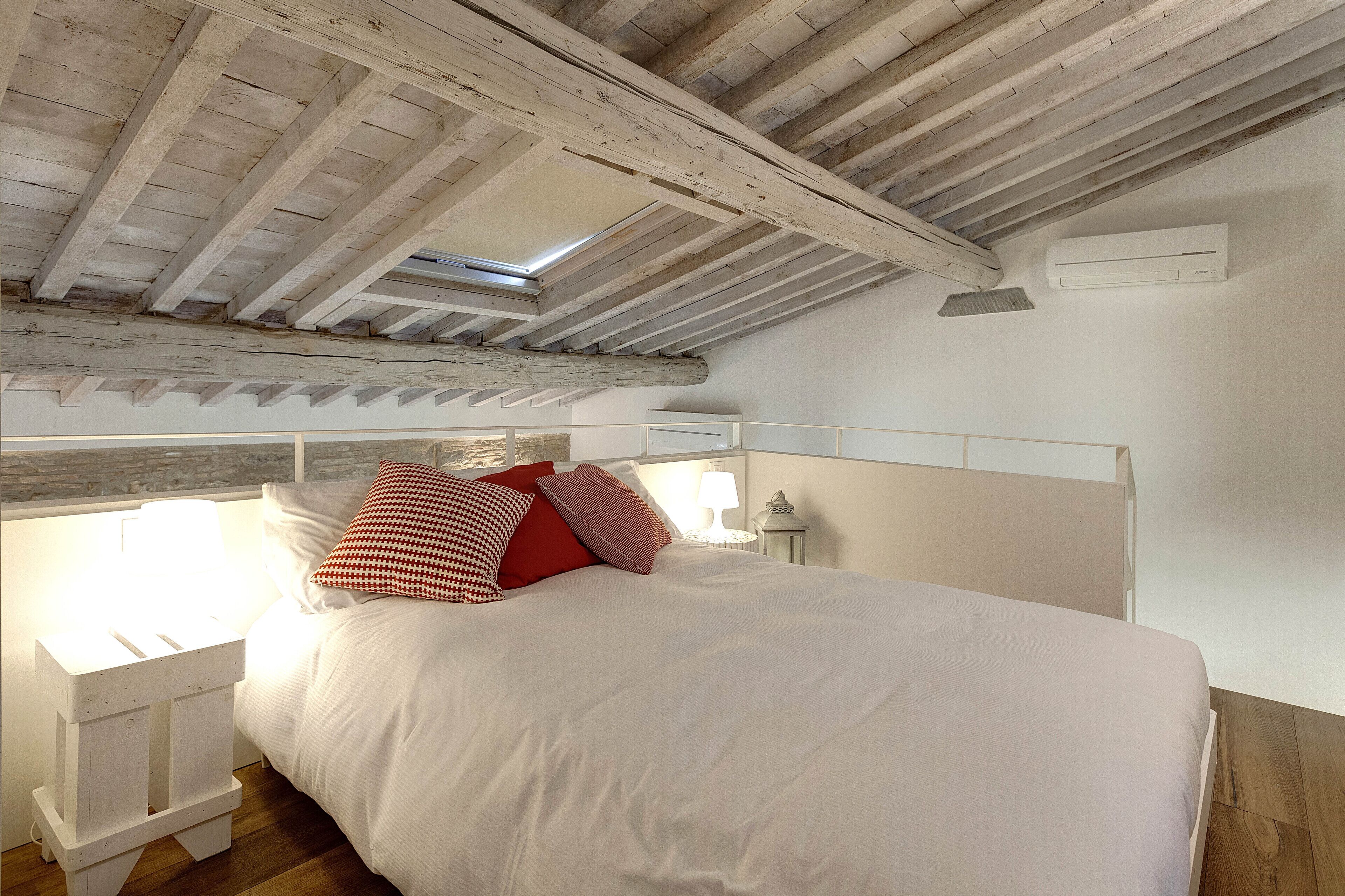 A bedroom with a wooden ceiling, a skylight, a bed with white bedding and red pillows, a bedside table with a lamp, and an air conditioning unit on the wall.