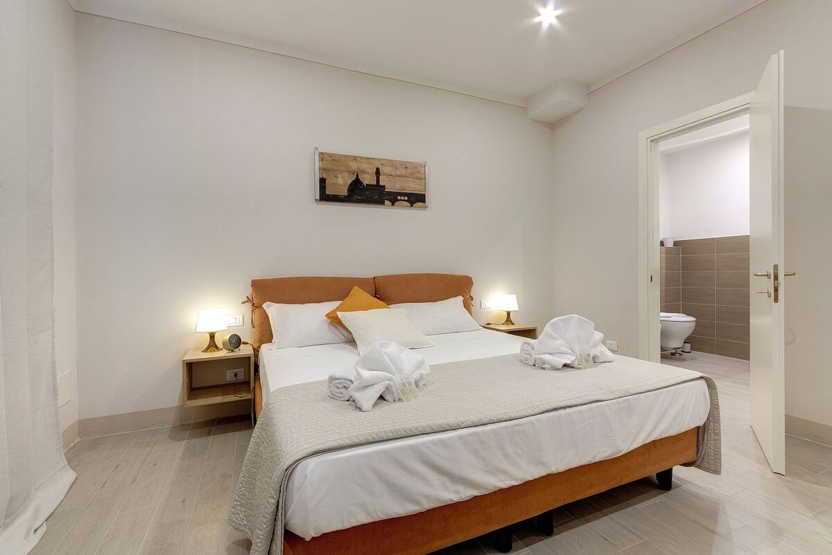 A neatly arranged hotel room with a large bed, two bedside tables with lamps, a wall-mounted artwork, and a door leading to a bathroom.
