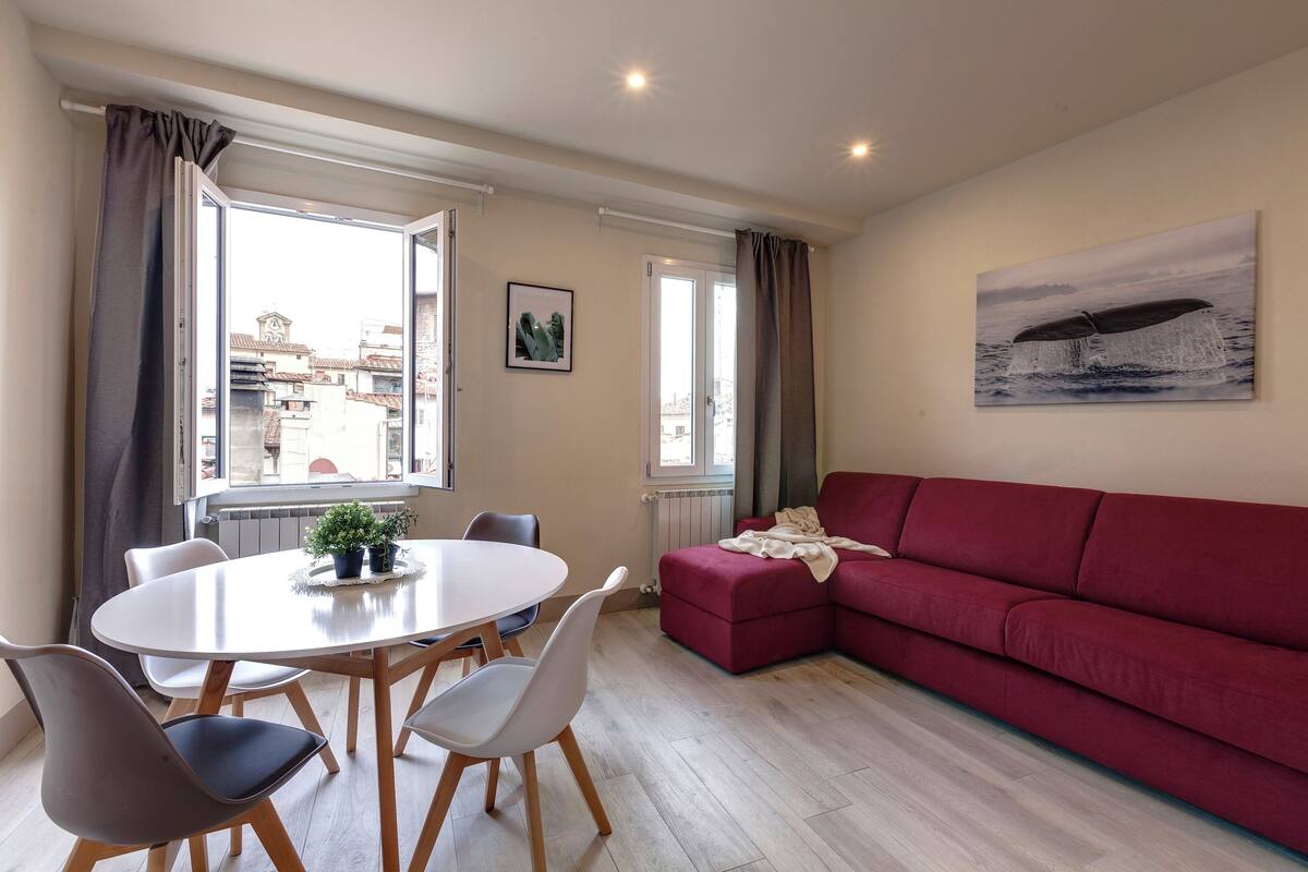 A modern living room with a red sofa, a dining table with chairs, and a large window with curtains.