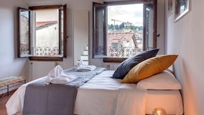 A bedroom with a bed, two open windows, a view of buildings, and a radiator. - Mamo Florence – Neri22 Apartments (Florence)