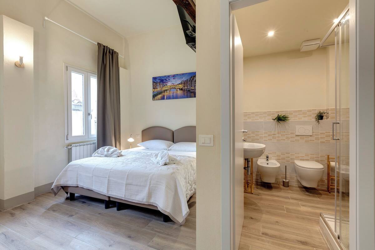 A bedroom with a bed, a window with curtains, a painting on the wall, and a bathroom with a toilet, bidet, and sink.