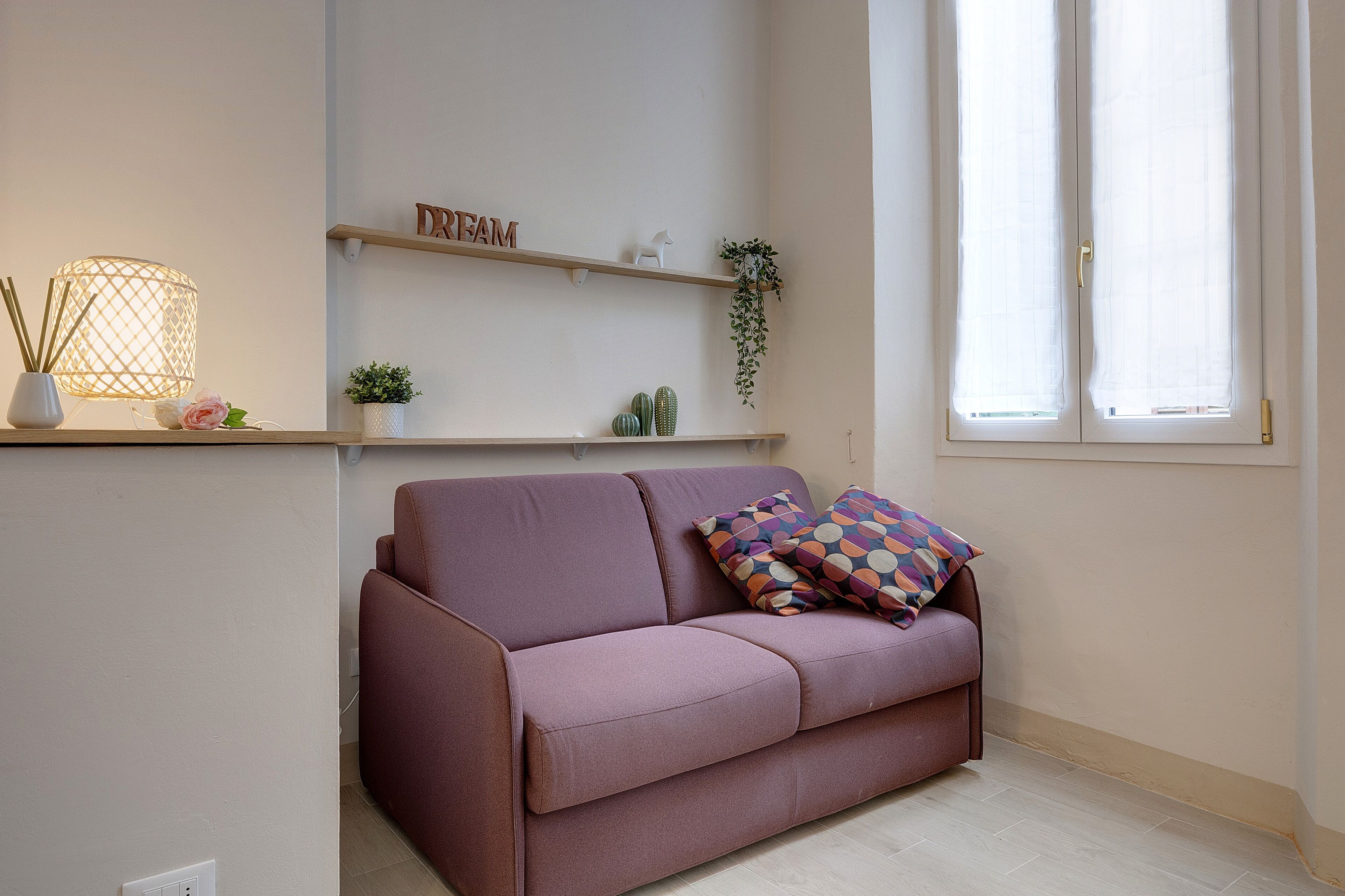 Un salon moderne avec un canapé violet, une lampe décorative, une étagère avec des plantes et un panneau indiquant le mot « Rêve », ainsi qu’une fenêtre ornée de rideaux légers.
