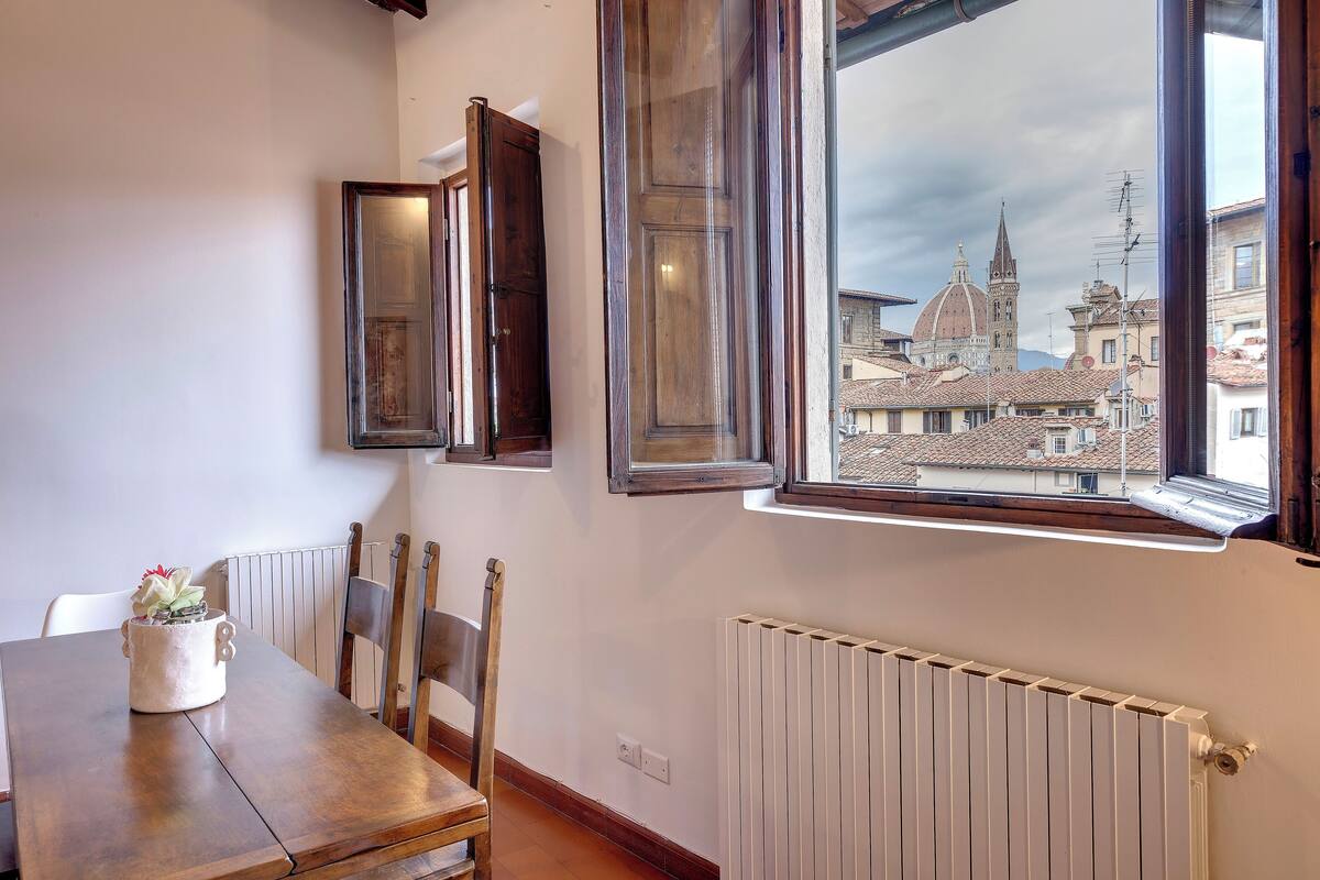 A dining area with a wooden table, chairs, a white radiator, and a window with a view of a historic building.