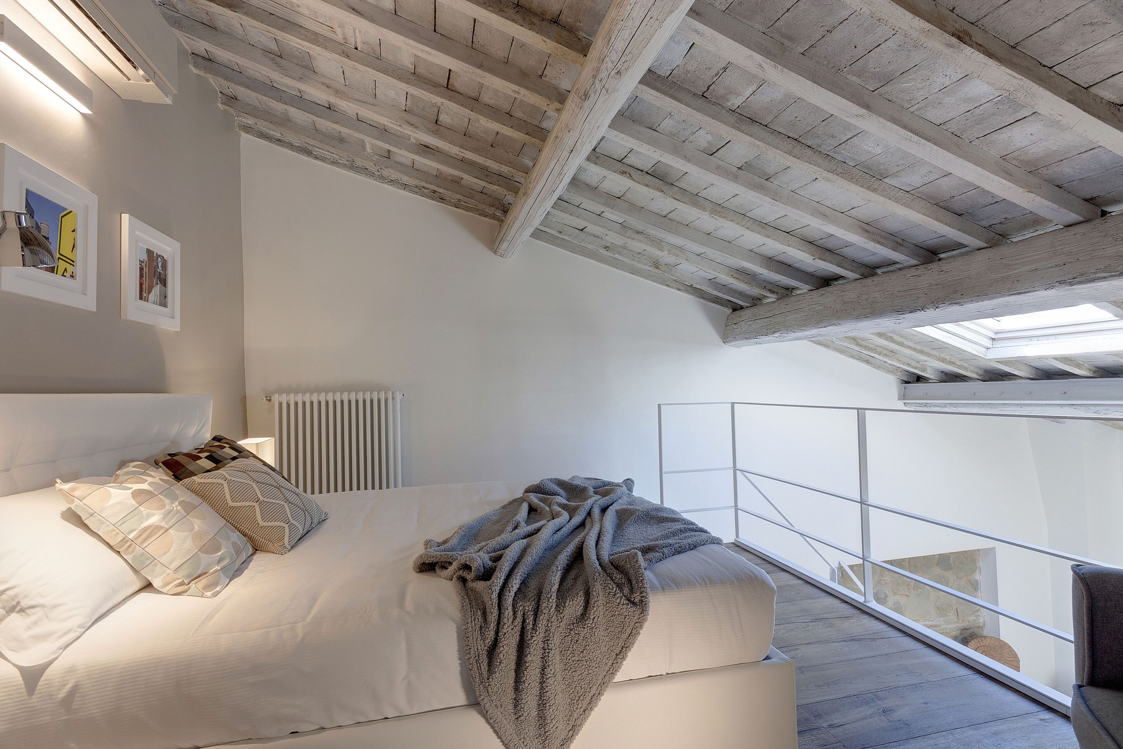 A bedroom with a high ceiling, exposed wooden beams, a bed with white bedding, and a view of a balcony.