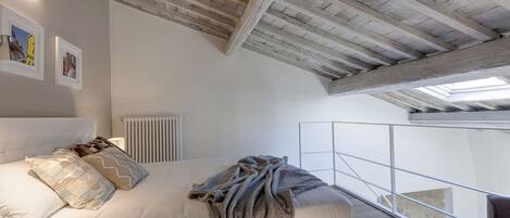 A bedroom with a high ceiling, exposed wooden beams, a bed with white bedding, and a view of a balcony.