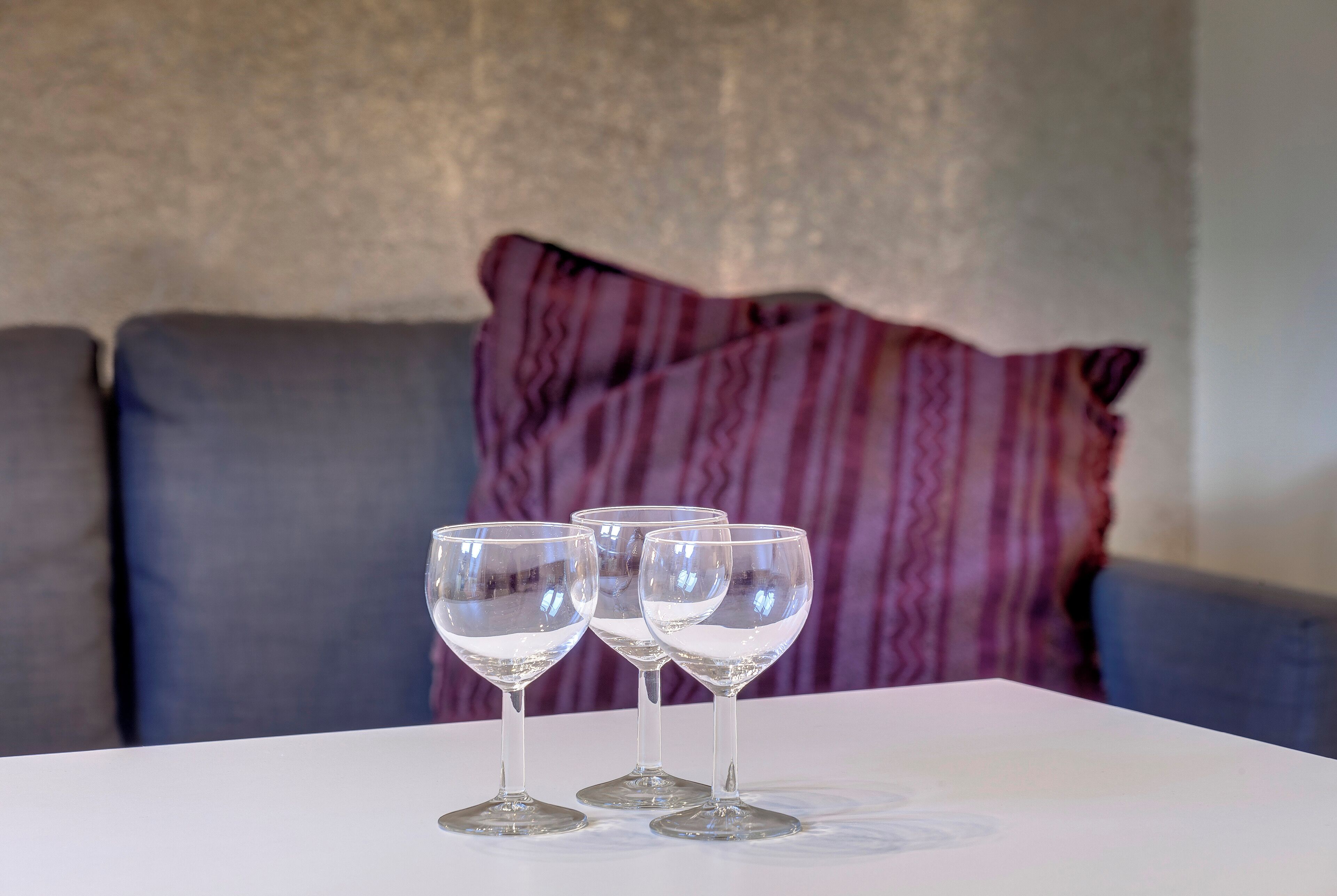 Three wine glasses on a white table in a room with a grey couch and a purple striped pillow in the background.