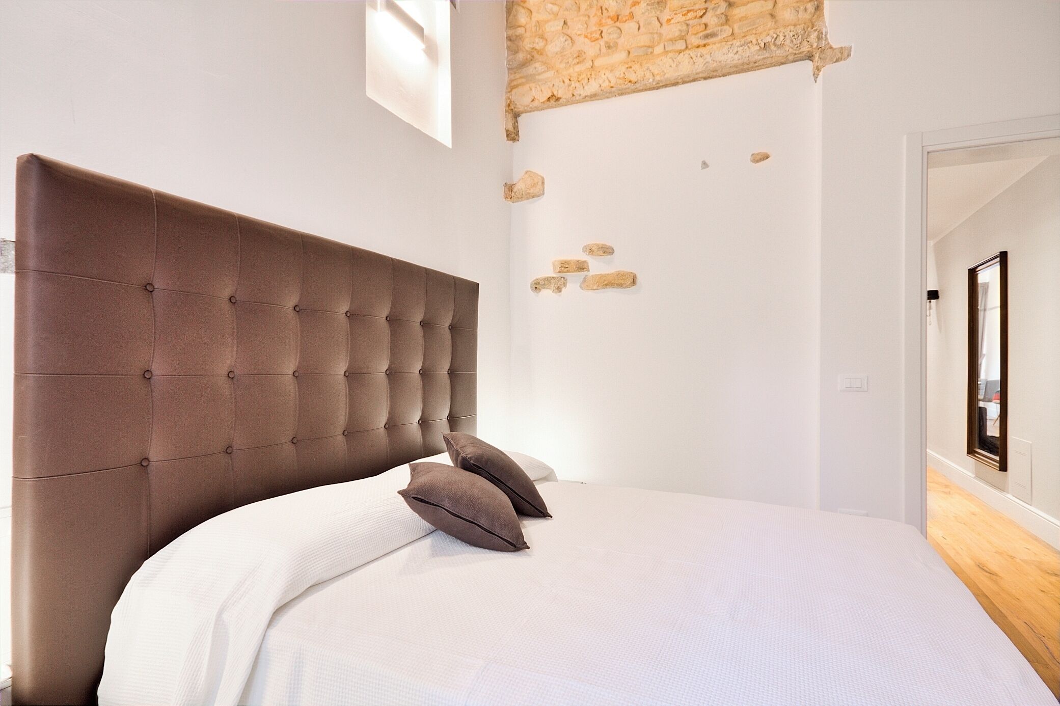 A bedroom with a tufted headboard, a white bedspread, and a stone accent wall.
