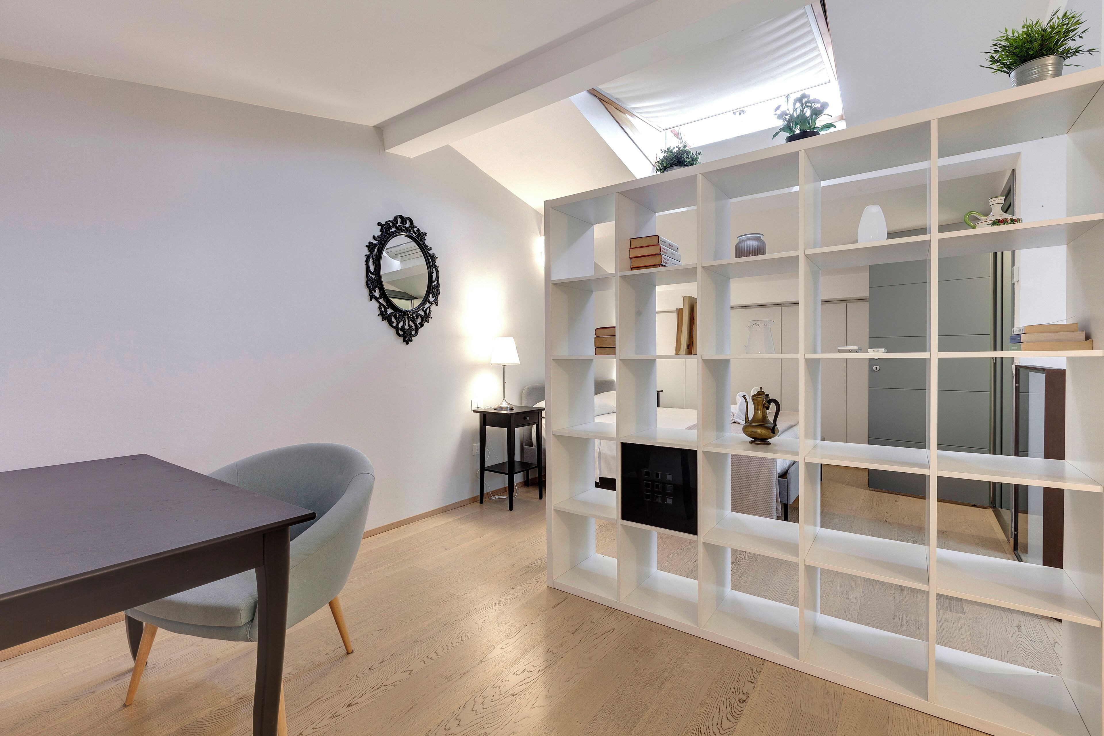 A modern room with a dining table, a light blue chair, a white shelving unit, and a small table with a lamp.