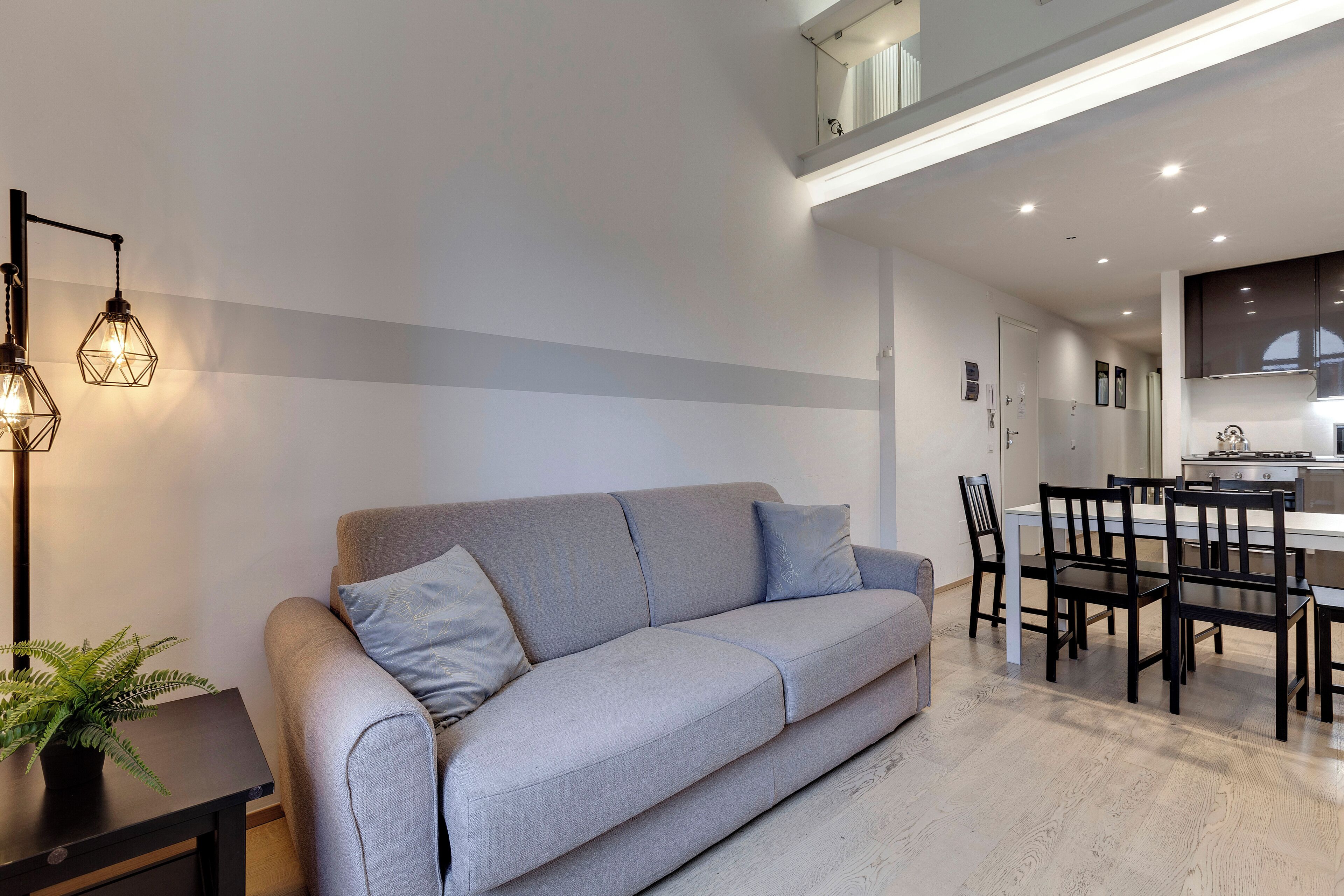 A modern living room with a grey sofa, a dining table with chairs, and a kitchen in the background.