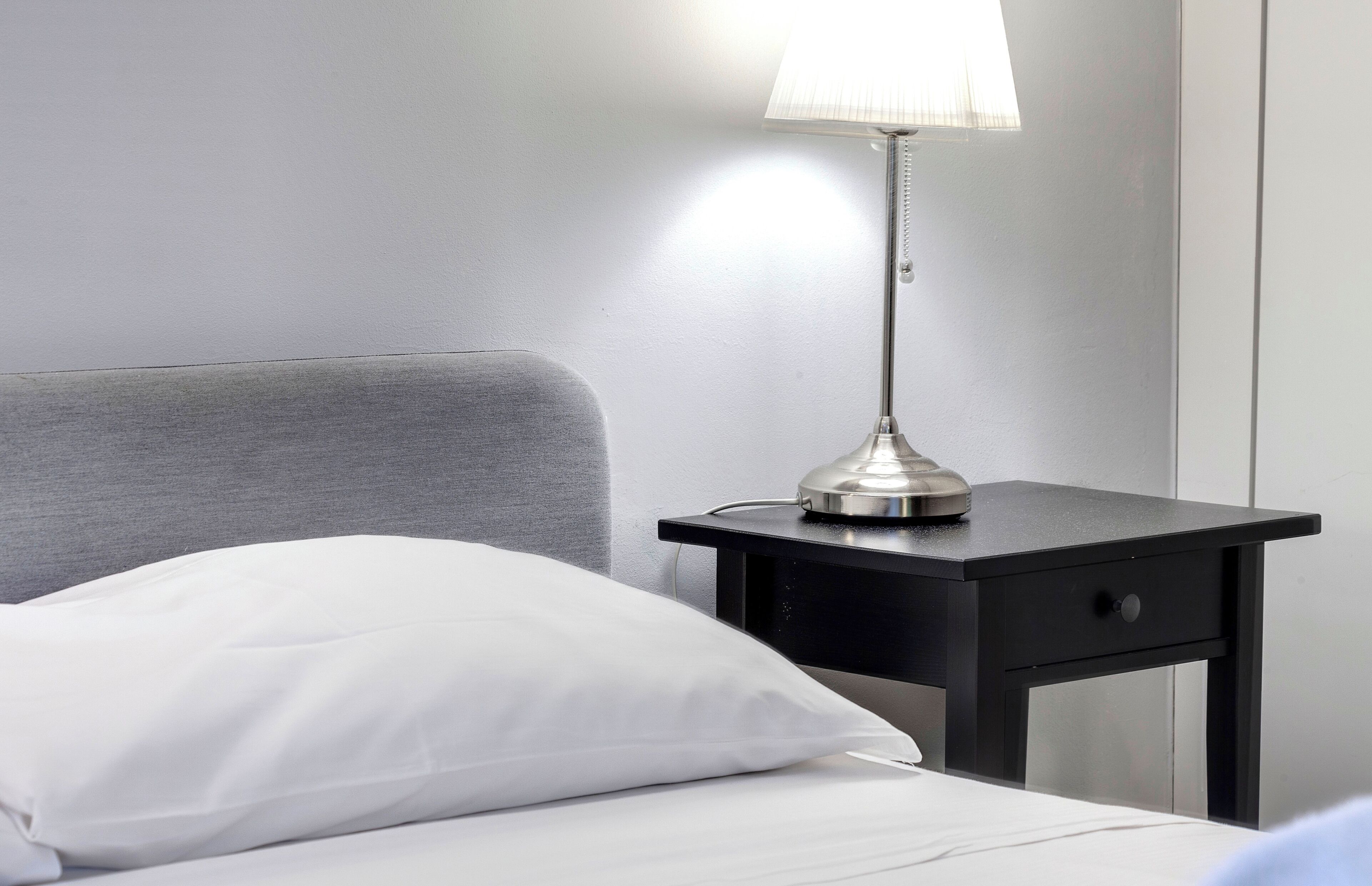 A neatly made bed with a white pillow and a black nightstand beside it. A modern table lamp with a metallic base and a white lampshade is placed on the nightstand.