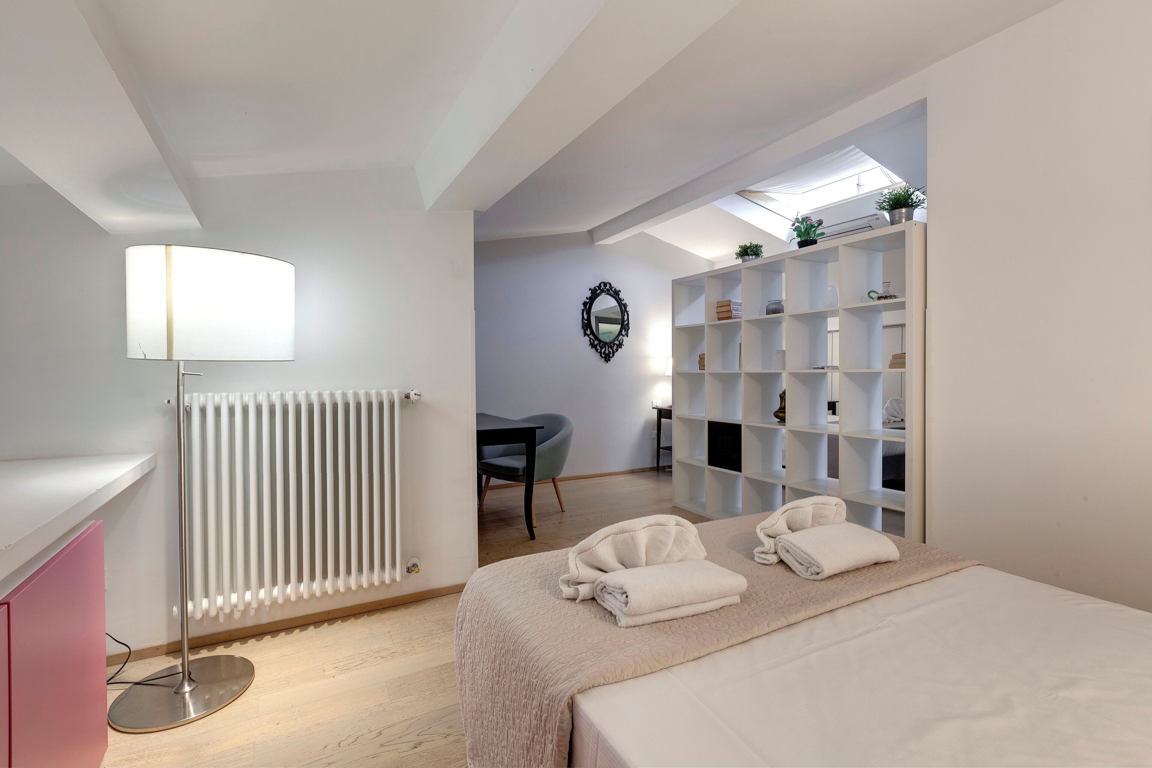 A modern hotel room with a bed, a desk, a chair, a radiator, and a white shelving unit with plants.