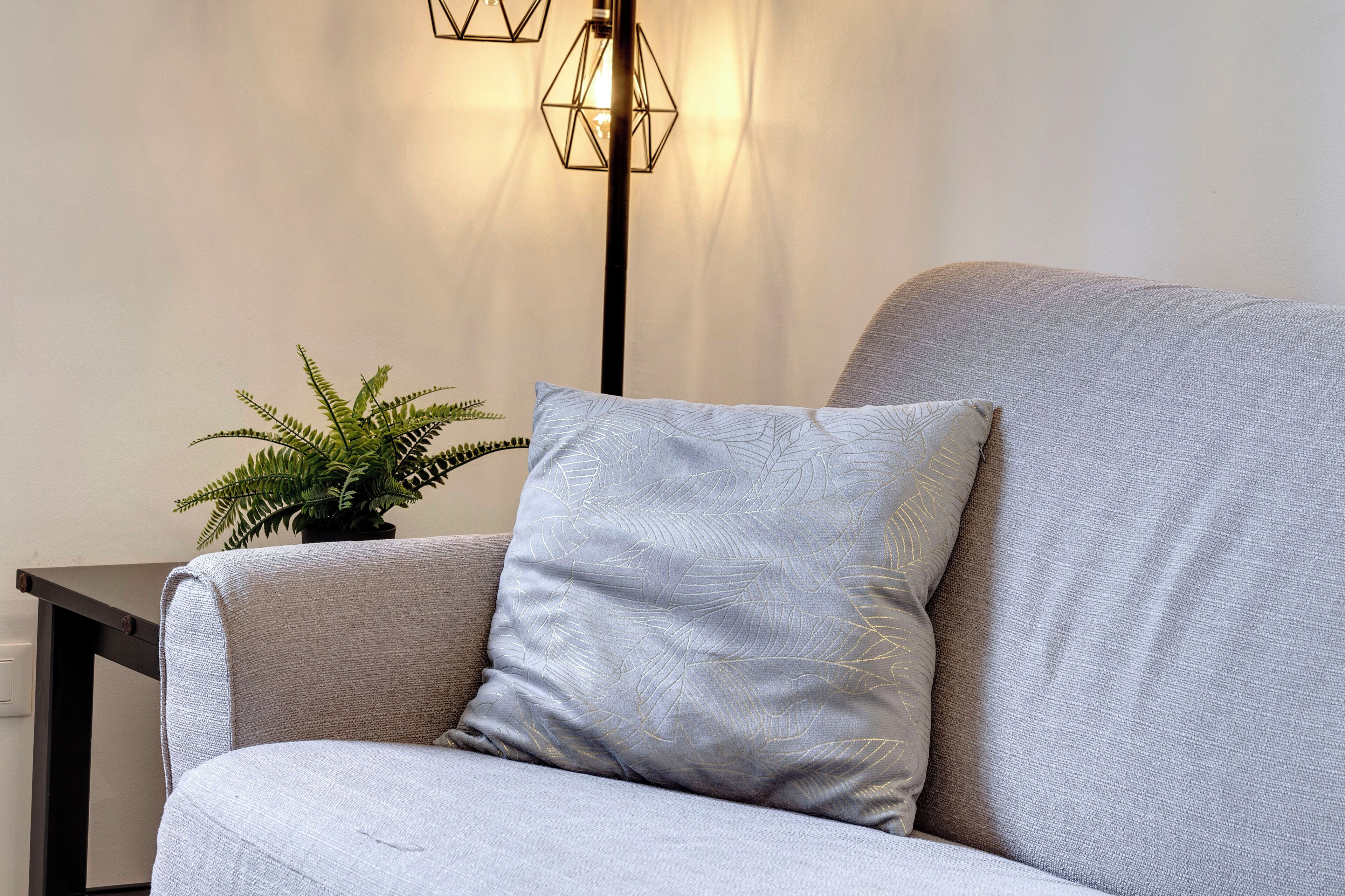 A modern living room with a grey sofa, a patterned pillow, a small potted plant, and a geometric floor lamp.