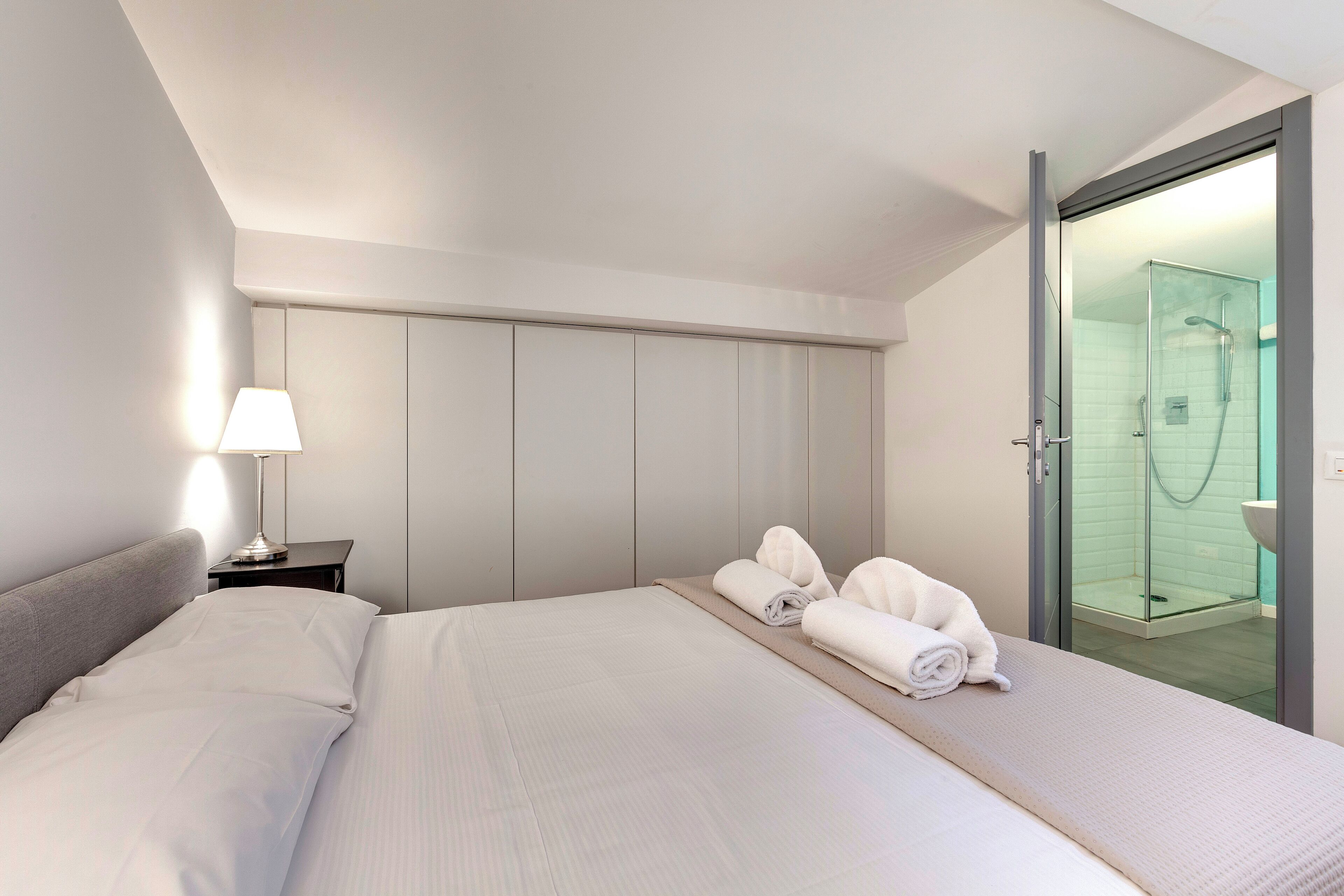A modern hotel room with a large bed, a bedside table with a lamp, and a glass-enclosed shower area.