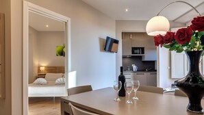 Ein modernes Hotelzimmer mit Essbereich, einem an der Wand befestigten Fernseher, einer Küche im Hintergrund und einer Vase mit roten Rosen auf dem Esstisch.