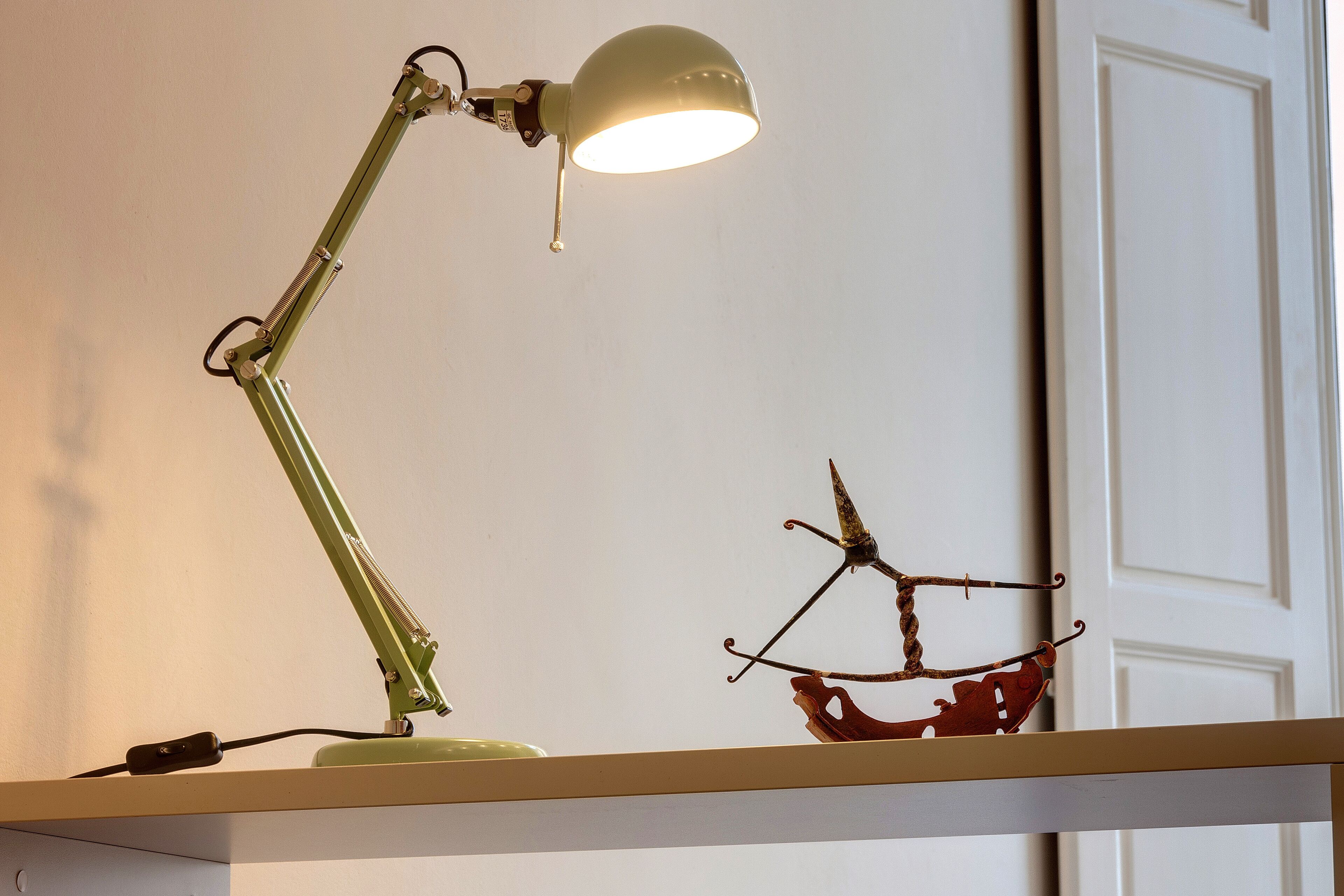 A desk lamp with a flexible neck and a white shade, positioned on a shelf.