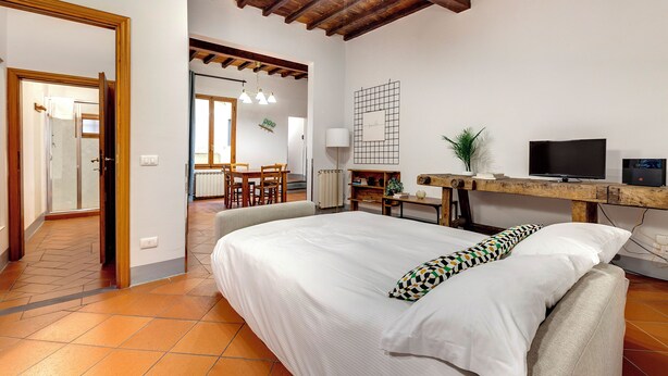 A bedroom with a large bed, a desk with a computer, a TV, and a wooden ceiling. - Mamo Florence - Zeno Apartment (Florence)